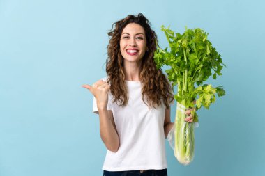 Mavi arka planda izole edilmiş bir kereviz tutan genç bir kadın bir ürünü sunmak için yan tarafı işaret ediyor.