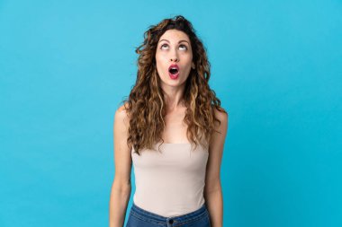 Genç beyaz kadın mavi arka planda izole edilmiş yukarı bakıyor ve şaşırmış bir ifadeyle