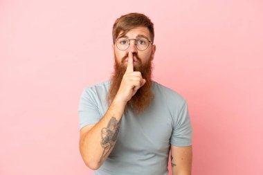 Pembe arka planda izole edilmiş genç, beyaz bir adam sessiz bir jest işareti gösteriyor. Parmağını ağzına sokuyor.