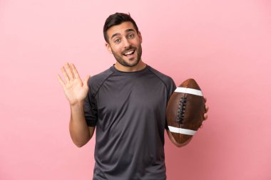 Genç beyaz adam rugby oynuyor. Pembe arka planda izole edilmiş. Mutlu bir ifadeyle selam veriyor.