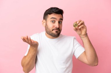 Pembe arka planda izole edilmiş Bitcoin tutan genç bir adam, ellerini kaldırırken şüphe duyuyor.