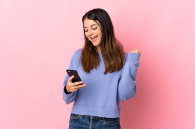 Pembe arka planda izole edilmiş genç beyaz kadın, telefon zafer pozisyonunda.