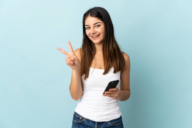 Mavi arka planda izole edilmiş cep telefonu kullanan genç beyaz kadın gülümsüyor ve zafer işareti gösteriyor.