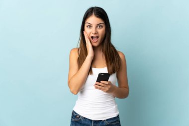 Mavi arka planda izole edilmiş, şaşırmış ve şok olmuş bir yüz ifadesiyle cep telefonu kullanan beyaz bir kadın.