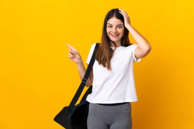 Sarı arka planda izole edilmiş spor çantalı genç bayan şaşırdı ve parmağını yana çevirdi.
