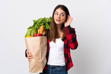 Beyaz arka planda izole edilmiş sebzeleri olan genç bir beyaz elini kulağına koyarak bir şeyler dinliyor.