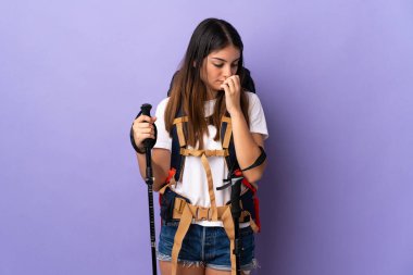 Sırt çantası ve yürüyüş direkleri olan genç bir kadın. Mor arka planda izole edilmiş. Şüpheleri var.