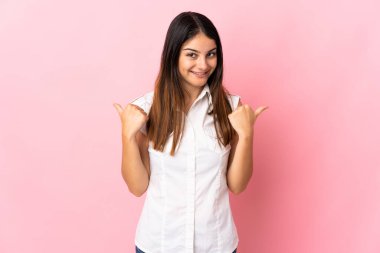 Pembe arka planda soyutlanmış genç beyaz bir kadın. Baş parmakları havada ve gülümsüyor.