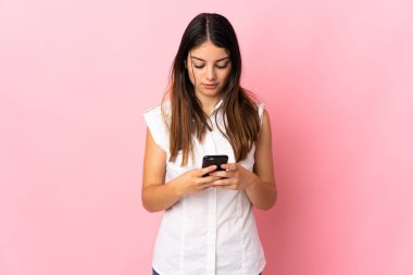 Pembe arka planda izole edilmiş genç beyaz kadın cep telefonuyla bir mesaj gönderiyor.
