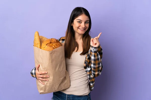 Молодая Женщина Держит Сумку Полную Хлеба Изолированы Фиолетовом Фоне Указывая — стоковое фото