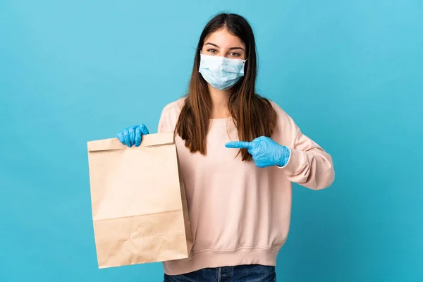 マスクでコロナウイルスから保護し 青の背景に隔離された食料品の買い物袋を保持し それを指す若い女性 — ストック写真