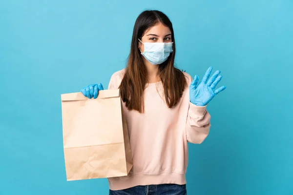 マスクでコロナウイルスから保護し 幸せな表情で手で塩漬け青の背景に隔離された食料品の買い物袋を保持している若い女性 — ストック写真