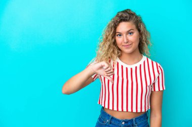 Mavi arka planda izole edilmiş kıvırcık saçlı kız olumsuz ifadeyle baş parmağını gösteriyor.