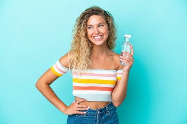 Mavi arka planda izole edilmiş su şişesi olan genç sarışın kadın kolları kalçasında ve gülümsüyor.