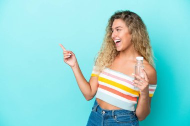 Mavi arka planda izole edilmiş su şişesi olan genç sarışın bir kadın parmak göstererek bir ürün sunuyor.