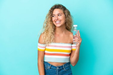 Mavi arka planda bir şişe suyu olan genç sarışın kadın yan tarafa bakıyor ve gülümsüyor.
