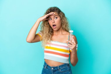 Mavi arka planda su şişesi olan genç sarışın kadın yan tarafa bakarken sürpriz jest yapıyor.