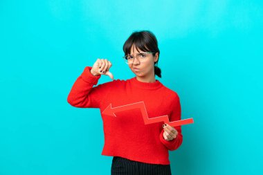 Mavi arka planda izole edilmiş genç bir kadın aşağıya doğru bir ok tutuyor ve kötü sinyal veriyor.