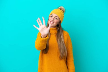 Kışlık ceket giyen genç beyaz kadın mavi arka planda izole edilmiş, parmaklarıyla beşi sayıyor.