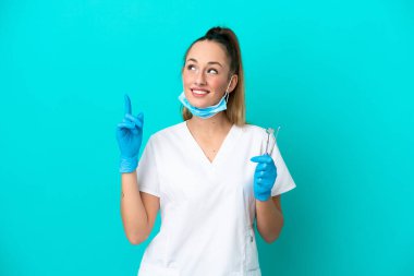 Elinde mavi arka planda izole edilmiş aletler tutan beyaz dişçi kadın harika bir fikre işaret ediyor.