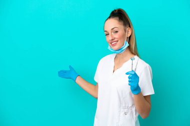 Beyaz dişçi kadın, elinde mavi arka planda izole edilmiş aletler tutuyor. Davet etmek için ellerini yana uzatıyor.