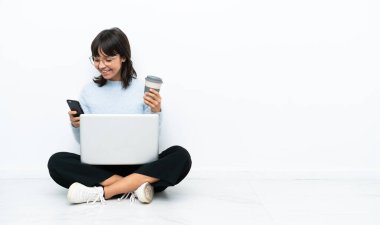 Genç melez kadın dizüstü bilgisayarla yerde oturuyor. Beyaz arka planda izole edilmiş. Elinde kahve ve cep telefonu var.