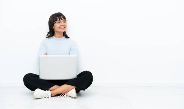 Genç melez kadın dizüstü bilgisayarla yerde oturuyor beyaz arka planda izole edilmiş, kolları bağlı ve mutlu.