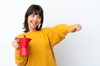 Beyaz arka planda kahve demliği tutan genç melez bir kadın baş parmak işareti yapıyor.