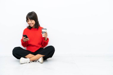 Genç melez bir kadın, beyaz arka planda izole bir şekilde yerde oturuyor. Elinde kahve ve cep telefonu var.