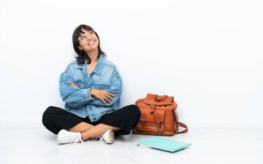 Genç bir kız öğrenci, beyaz arka planda tek başına oturmuş gülümserken yukarı bakıyor.