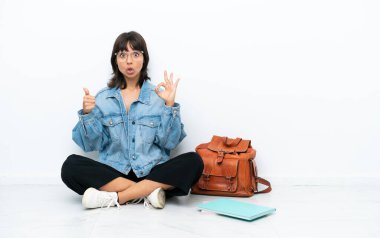 Genç bir bayan, beyaz arka planda tek başına oturuyor. Tamam işareti gösteriyor ve işaret veriyor.