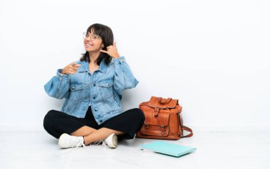 Genç bir kız öğrenci, beyaz arka planda tek başına oturuyor telefon hareketlerinde bulunuyor ve ön tarafı işaret ediyor.