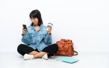 Genç bir kız öğrenci beyaz arka planda tek başına oturuyor. Elinde kahve ve cep telefonu var.