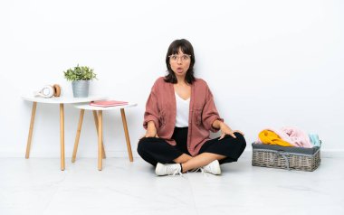 Genç melez kadın, katlanan giysiler, beyaz arka planda izole bir şekilde oturuyor ve yüzünde sürpriz bir ifade var.