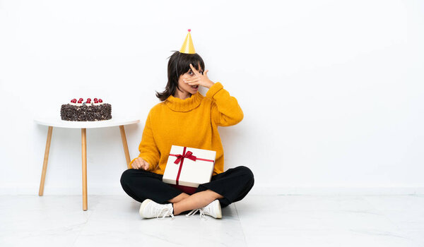 Young mixed race woman holding a gift sitting on the floor isolated on white background covering eyes by hands and smiling