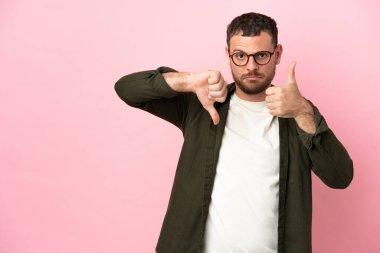 Pembe arka planda izole edilmiş genç Brezilyalı adam iyiye-kötüye işaret veriyor. Evet ya da hayır arasında kararsız