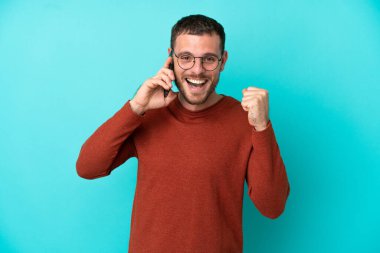 Mavi arka planda izole edilmiş cep telefonu kullanan genç Brezilyalı adam galibiyeti kutluyor.