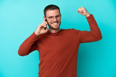 Mavi arka planda izole edilmiş cep telefonu kullanan Brezilyalı genç güçlü bir jest yapıyor.