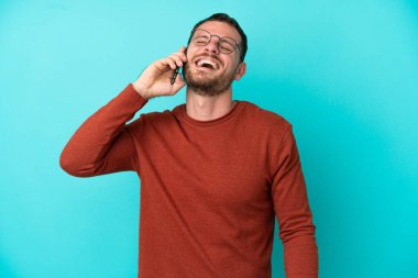 Mavi arka planda izole edilmiş cep telefonu kullanan genç Brezilyalı adam gülüyor.