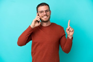 Mavi arka planda izole edilmiş cep telefonu kullanan genç Brezilyalı adam en iyinin işareti olarak parmağını kaldırıyor.