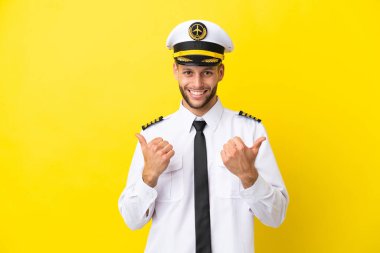 Kafkasyalı uçak pilotu sarı arka planda izole edilmiş. Baş parmakları havada ve gülümsüyor.