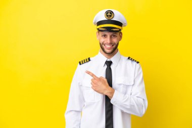 Kafkasyalı uçak pilotu sarı arka planda izole edilmiş bir ürün sunmak için yan tarafı işaret ediyor.