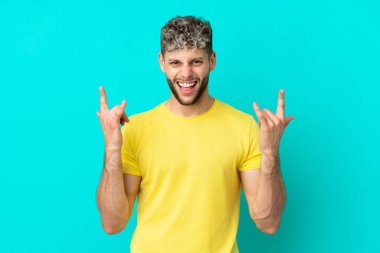 Mavi arka planda izole edilmiş yakışıklı beyaz adam boynuz hareketi yapıyor.