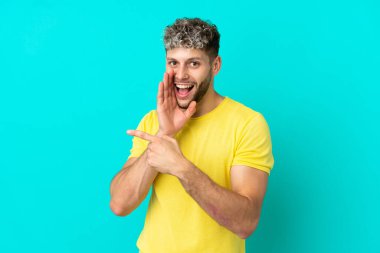 Mavi arka planda soyutlanmış yakışıklı beyaz bir adam bir ürün sunmak ve bir şeyler fısıldamak için kenarı işaret ediyor.