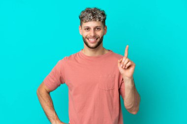 Mavi arka planda izole edilmiş yakışıklı beyaz adam en iyinin işareti olarak parmağını kaldırıyor.