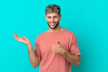 Genç, beyaz, mavi arka planda izole edilmiş bir adam. Kopyalama alanı hayali bir şey tutuyor. Bir ilan vermek için.