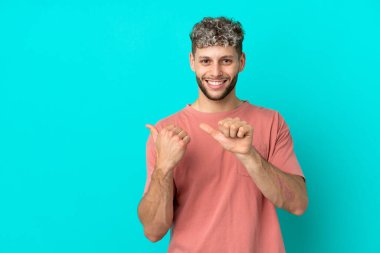 Mavi arka planda izole edilmiş yakışıklı beyaz adam bir ürünü sunmak için kenarı işaret ediyor.