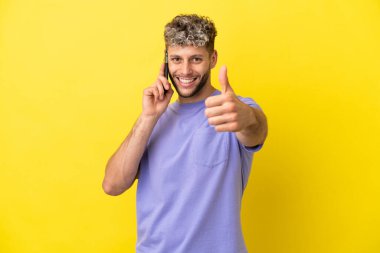 Sarı arka planda izole edilmiş cep telefonu kullanan beyaz bir genç. Başparmakları havada. Çünkü iyi bir şey oldu.