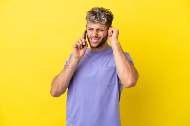 Sarı arka planda izole edilmiş cep telefonu kullanan genç beyaz adam hayal kırıklığına uğramış ve kulaklarını kapatmış.
