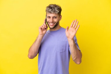 Sarı arka planda izole edilmiş cep telefonu kullanan beyaz bir adam mutlu bir ifadeyle selamlıyor.
